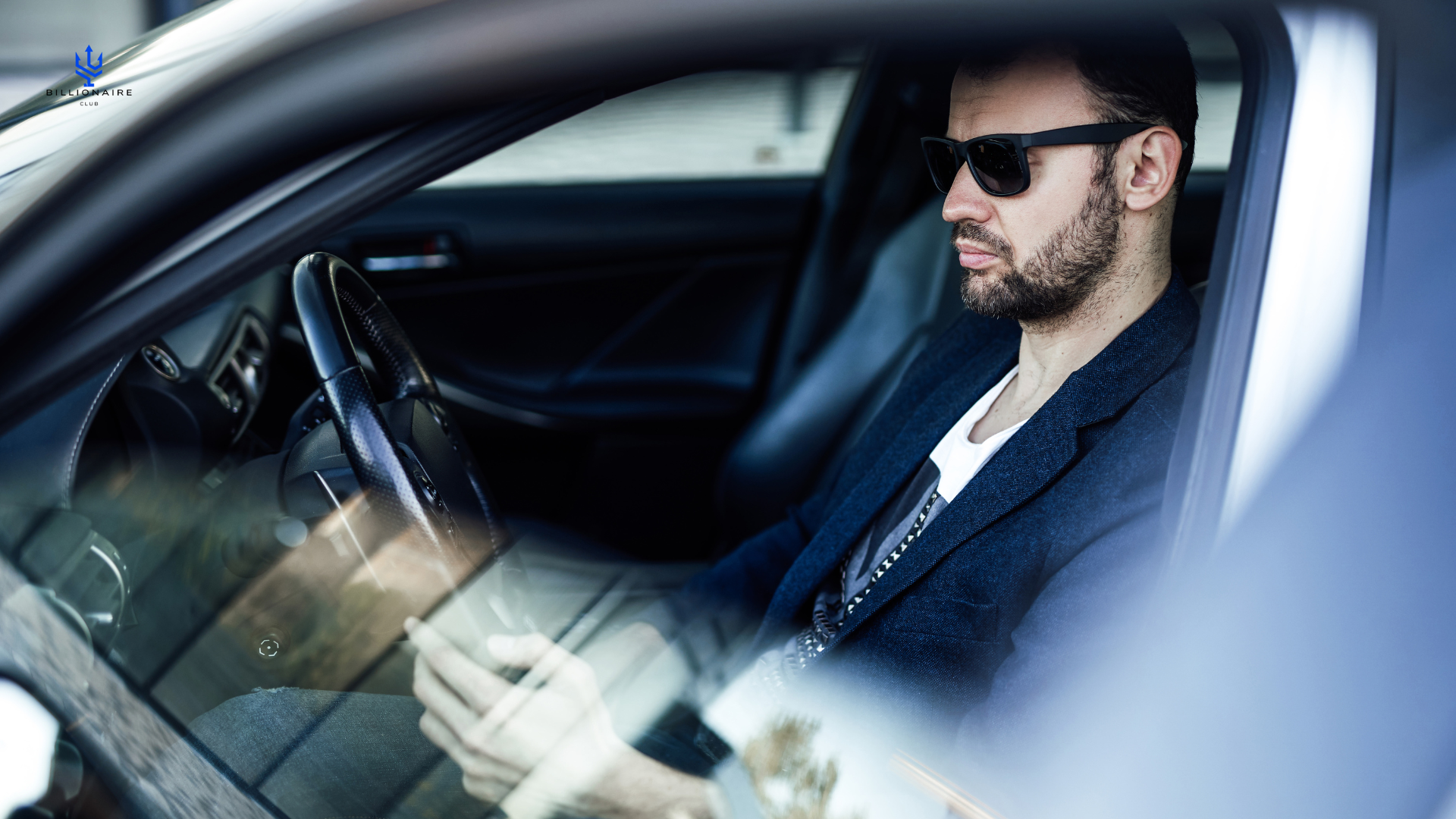 Man sitting in a car wearing sunglasses and a dark jacket.